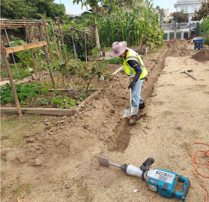 Landscaper wearing a safety vest digging a shallow trench with a shovel in a large, dry garden plot, with a blue jackhammer resting in the foreground.