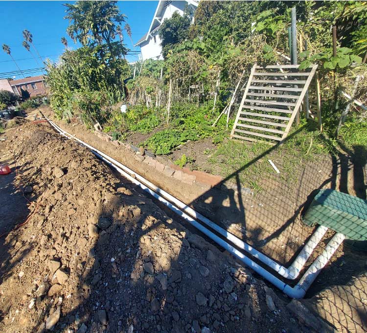 Long trench dug in backyard garden soil with white PVC irrigation pipes laid out along a chain-link fence line near a green valve box.