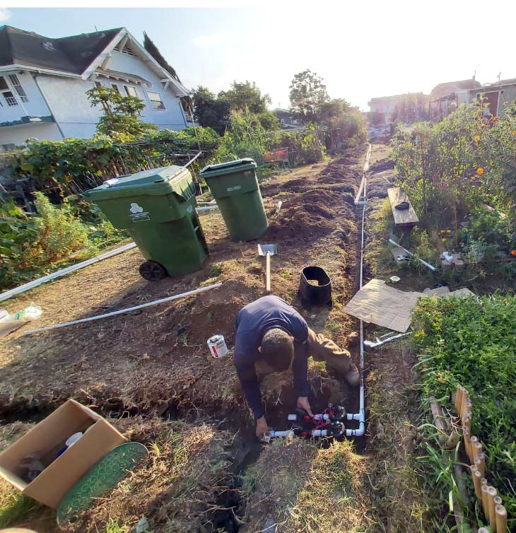 Installer kneeling in a dirt trench connecting a sprinkler valve manifold system with PVC pipes in a residential backyard garden setting for irrigation installation