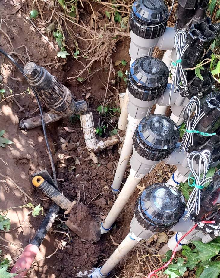 Close-up view of a multi-zone wired sprinkler valve manifold connected to white PVC pipes routed into a hole in the ground.