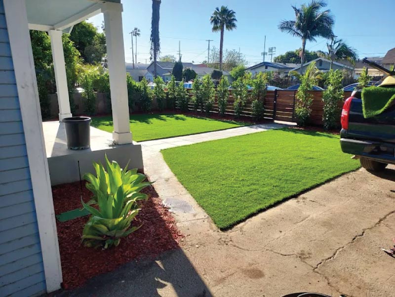 A manicured residential backyard featuring a vibrant, level lawn, a wooden privacy fence, and drought-tolerant perimeter plantings with red mulch. 