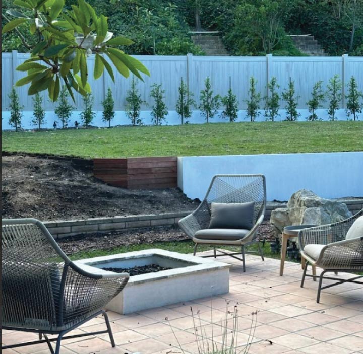 A modern outdoor living space featuring a stone patio with a fire pit, stylish wicker lounge chairs, and a clean, multi-level lawn design.